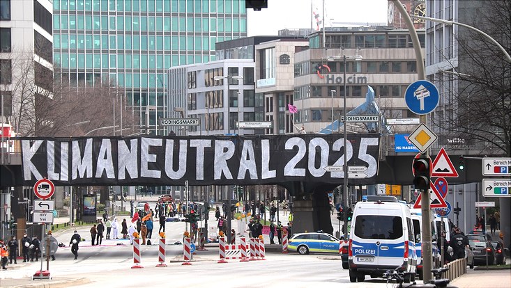 Straßensperrung wegen Klimademonstration in Hamburg, ein großes Banner mit der Aufschrift "Klimaneutral 2025" hängt über die ganze Breite einer Fußgängerbrücke über der Straße.