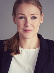 Portrait von Cathrin Despotovic. Cathrin trägt eine weißes Oberteil und einen schwarzen Blazer. Cathrins Haare sind dunkelblond und zu einem Pferdeschwanz gebunden, der Cathrin über die Schulter fällt. Cathrin lächelt. Der Hintergrund ist grau.
